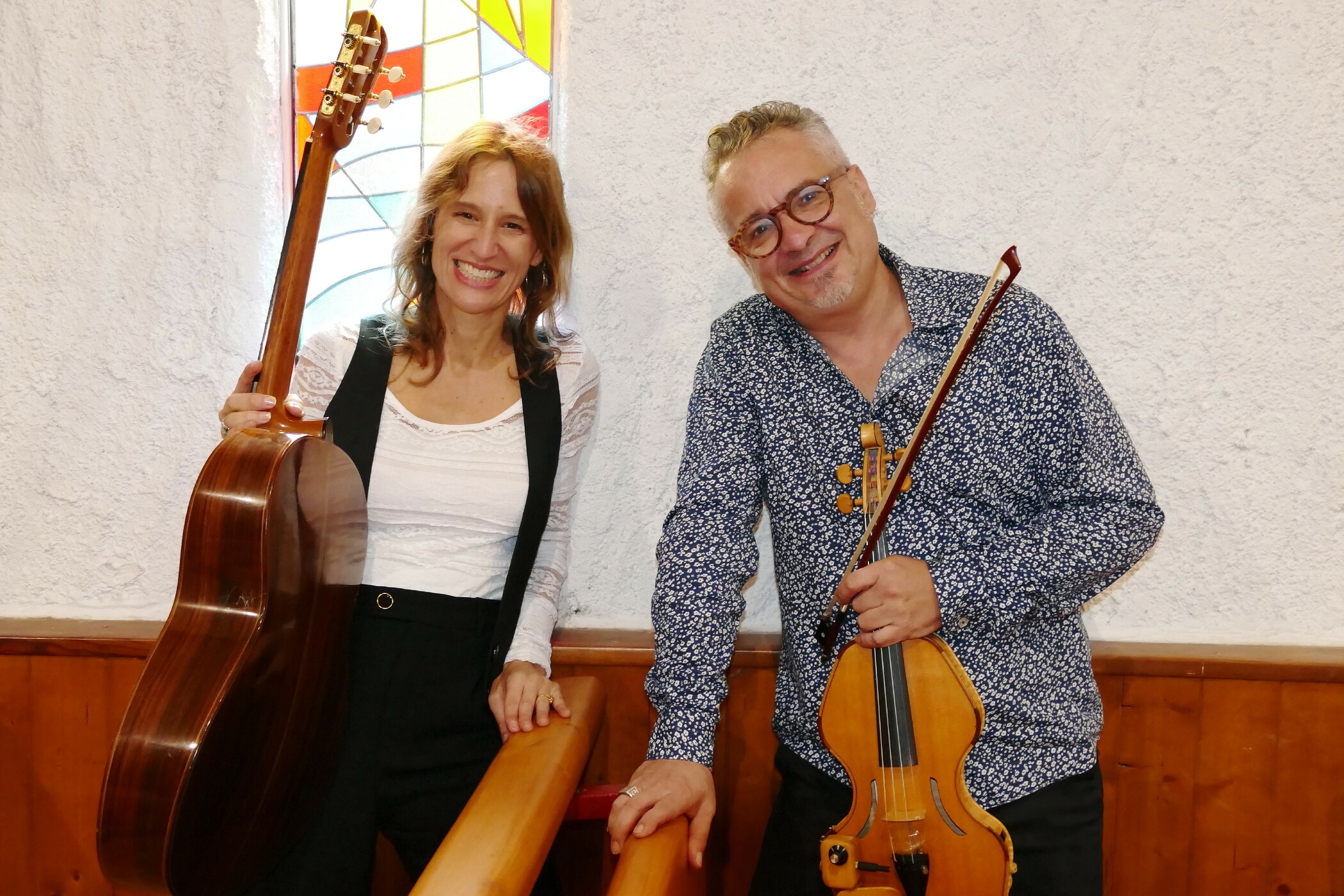 Une guitariste et un violoncelliste debout, dans une église et devant un vitrail coloré. Ils tiennent leur instrument d’une main et sourient.